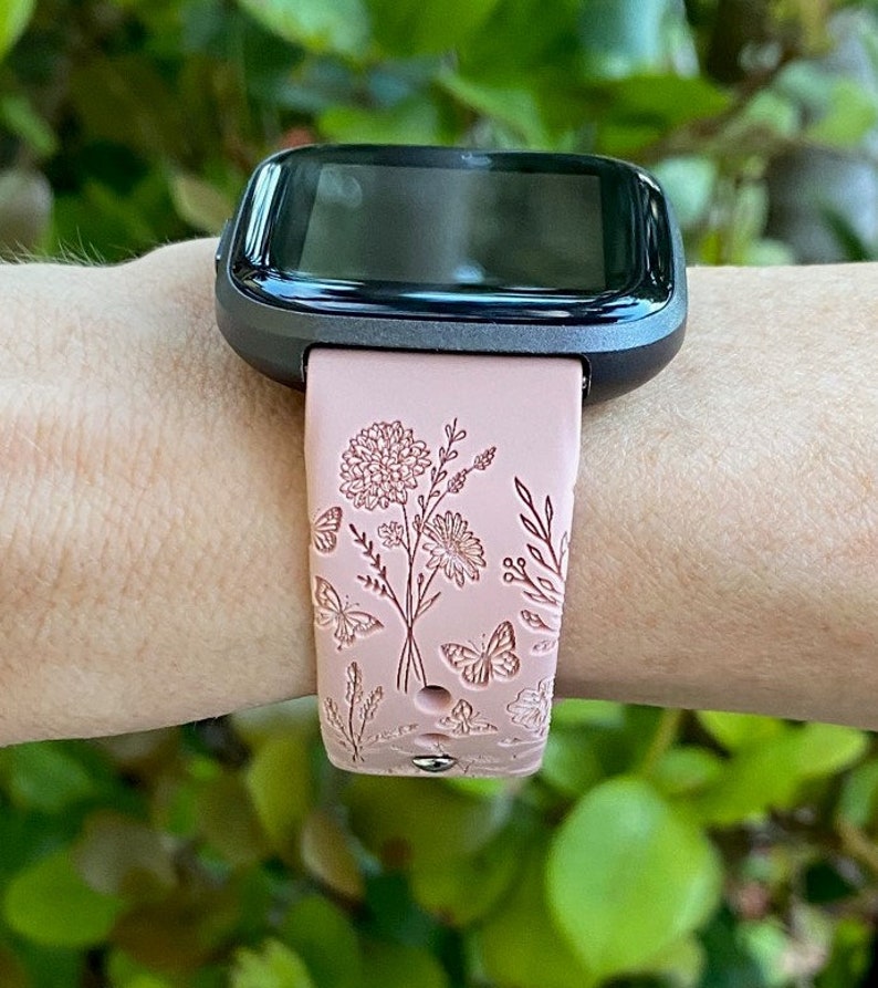 May include: A pink silicone watch band with a floral and butterfly design. The band is compatible with a Fitbit watch.