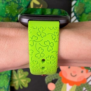 May include: A green smartwatch band with a clover pattern. The band is made of silicone and has a black watch face.