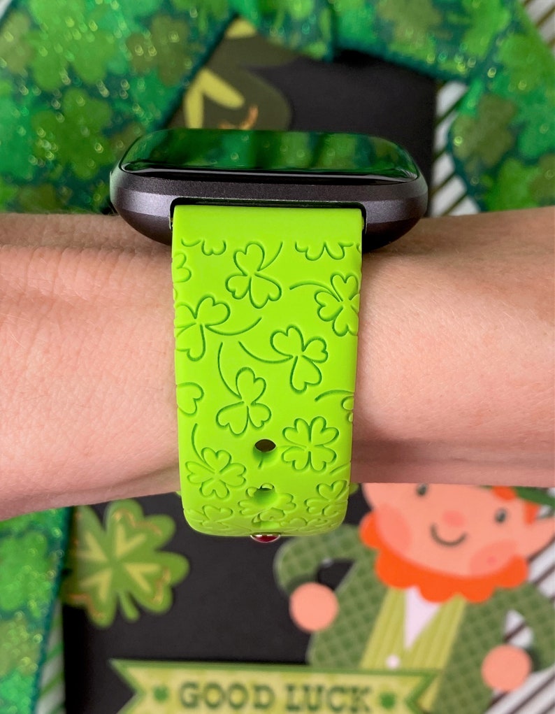 May include: A green smartwatch band with a clover pattern. The band is made of silicone and has a black watch face.