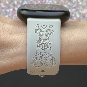 K&ouml;nnte beinhalten: Hellgraues Uhrenarmband mit einem Schnauzer-Hund-Design und drei Herzen &uuml;ber dem Kopf des Hundes. Das Armband ist an einer schwarzen Smartwatch befestigt. Das Hundedesign ist in das Armband ge&auml;tzt.