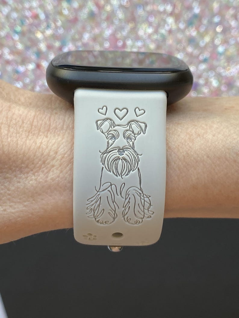 K&ouml;nnte beinhalten: Hellgraues Uhrenarmband mit einem Schnauzer-Hund-Design und drei Herzen &uuml;ber dem Kopf des Hundes. Das Armband ist an einer schwarzen Smartwatch befestigt. Das Hundedesign ist in das Armband ge&auml;tzt.