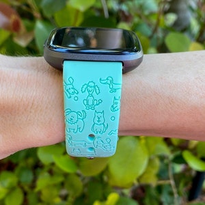 K&ouml;nnte beinhalten: Ein blaugr&uuml;nes Armband f&uuml;r eine Smartwatch mit schwarzem Zifferblatt. Das Armband hat ein sich wiederholendes Muster aus verschiedenen Hunderassen.