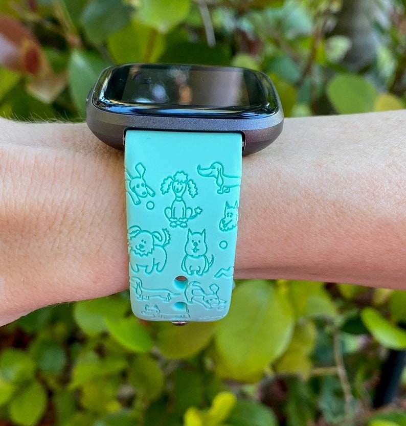 K&ouml;nnte beinhalten: Ein blaugr&uuml;nes Armband f&uuml;r eine Smartwatch mit schwarzem Zifferblatt. Das Armband hat ein sich wiederholendes Muster aus verschiedenen Hunderassen.