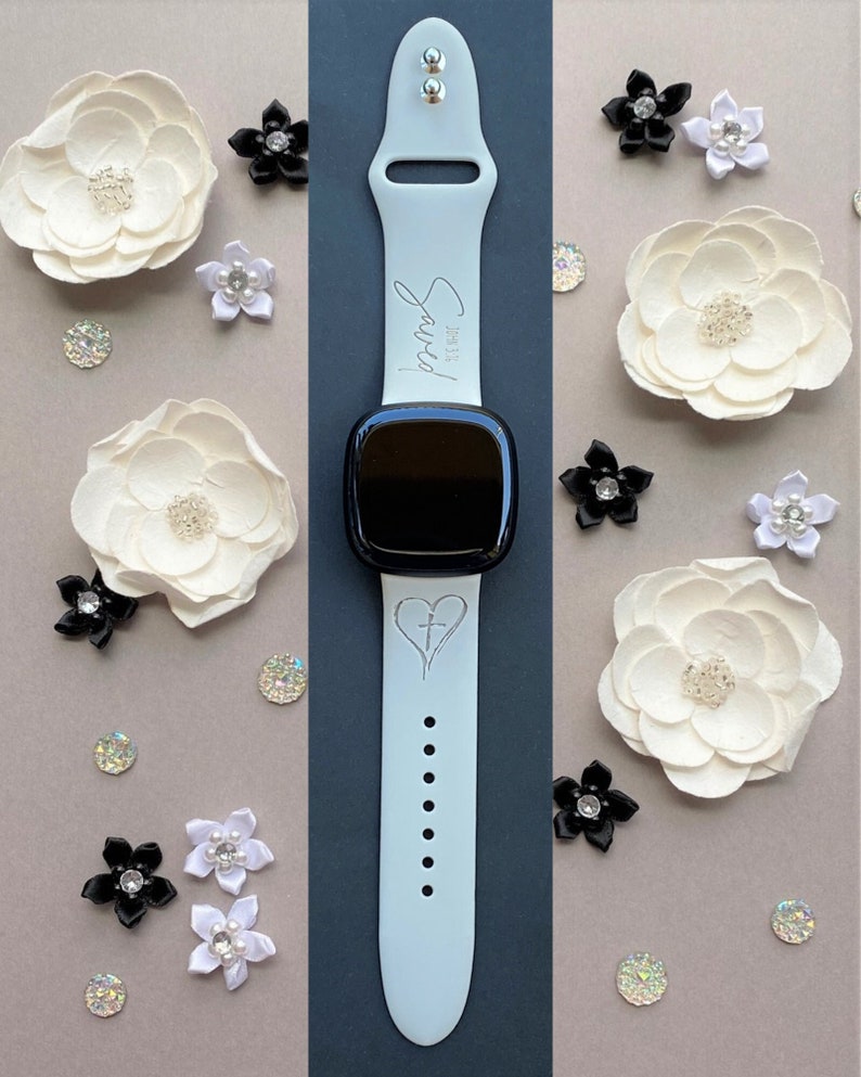 May include: A white silicone watch band with a black watch face. The band has a cross and heart design and the text "Sarah" is engraved on the band.
