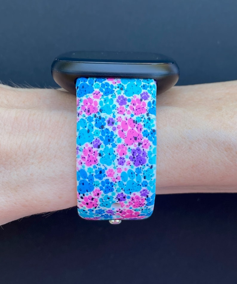Pu&ograve; includere: Un cinturino per orologio bianco con un motivo a forma di impronta di zampa in blu, rosa e viola. Il cinturino &egrave; realizzato in silicone ed &egrave; compatibile con la maggior parte degli smartwatch.