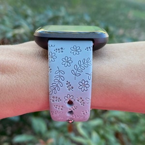 K&ouml;nnte beinhalten: Ein graues und wei&szlig;es Armband mit floralem Muster. Das Armband ist aus Silikon und hat eine silberne Schnalle.