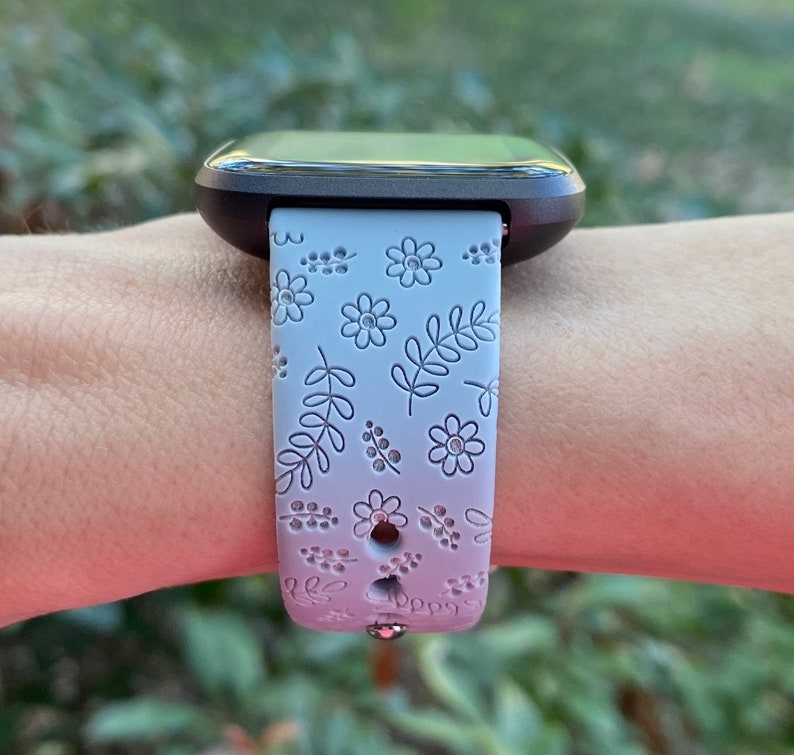 K&ouml;nnte beinhalten: Ein graues und wei&szlig;es Armband mit floralem Muster. Das Armband ist aus Silikon und hat eine silberne Schnalle.