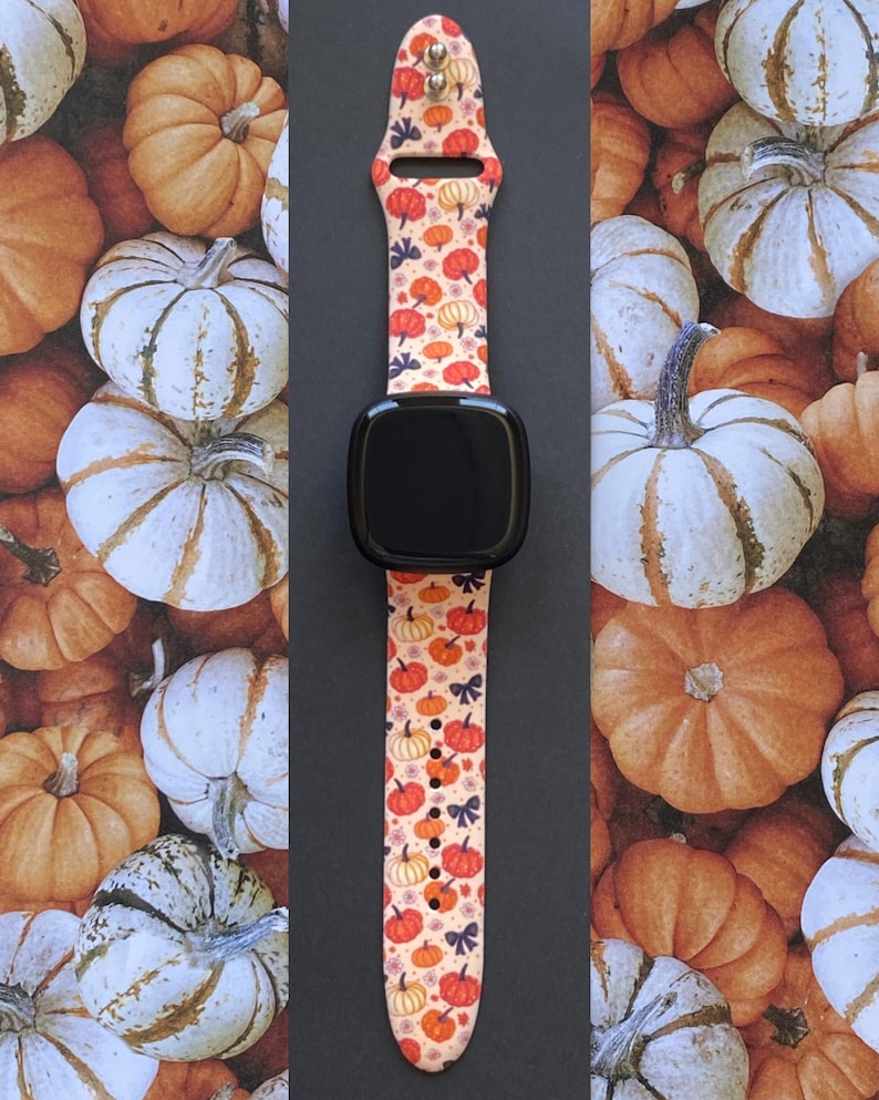 Może przedstawiać: Czarny smartwatch z paskiem we wz&oacute;r z pomarańczowymi, czerwonymi i ż&oacute;łtymi dyniami oraz małymi niebieskimi kokardkami na kremowym tle. Zegarek jest ustawiony na tle r&oacute;żnych dyni.