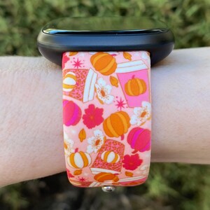 May include: A pink and orange patterned watch band with a design of pumpkins, coffee cups, and flowers. The band is made of silicone and is compatible with smartwatches.