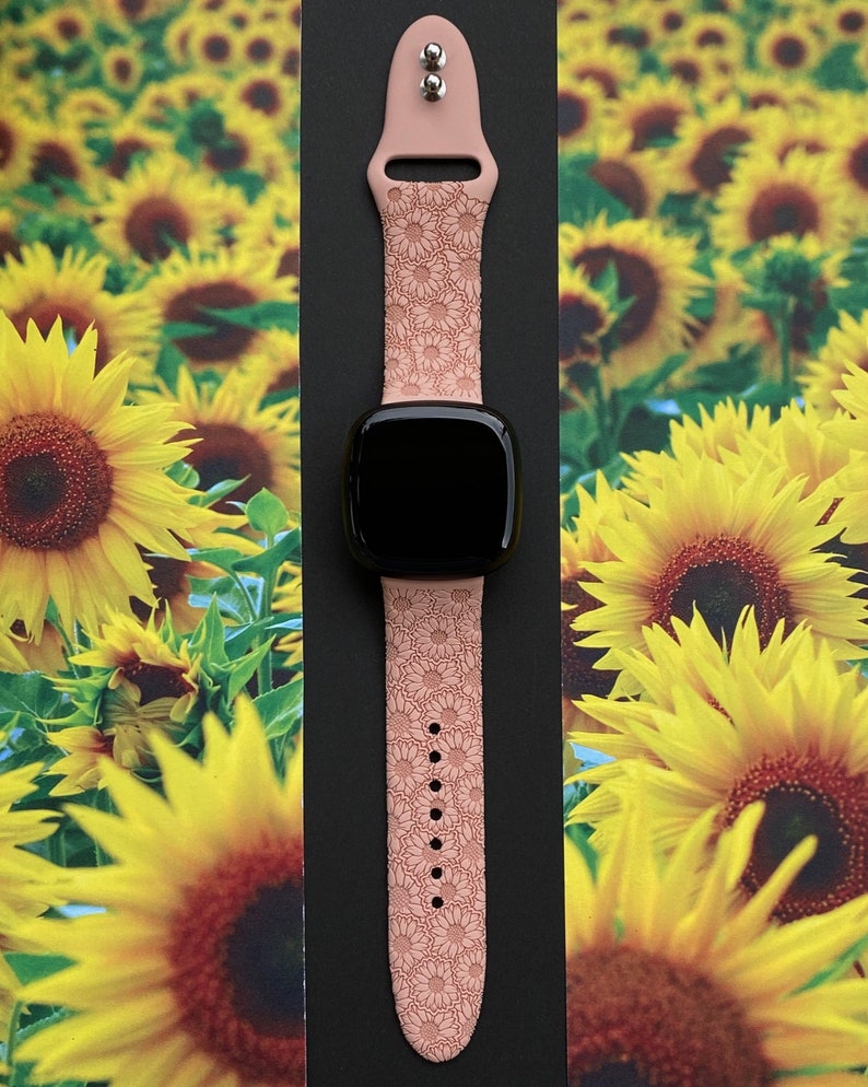 May include: A pink silicone watch band with a sunflower pattern. The band is designed for a smartwatch and has a black watch face.
