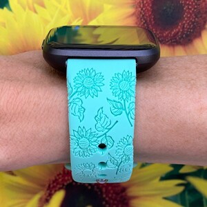 May include: A turquoise smartwatch band with a sunflower pattern. The band is made of silicone and has a textured surface.