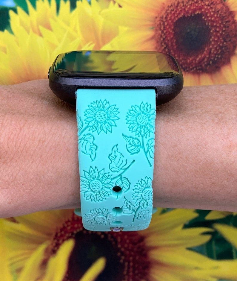 May include: A turquoise smartwatch band with a sunflower pattern. The band is made of silicone and has a textured surface.