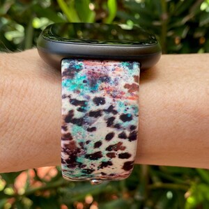 Może przedstawiać: Czarny smartwatch z kolorowym paskiem o abstrakcyjnym wzorze. Pasek ma białe tło z brązowymi, niebieskimi i turkusowymi plamami i kropkami.