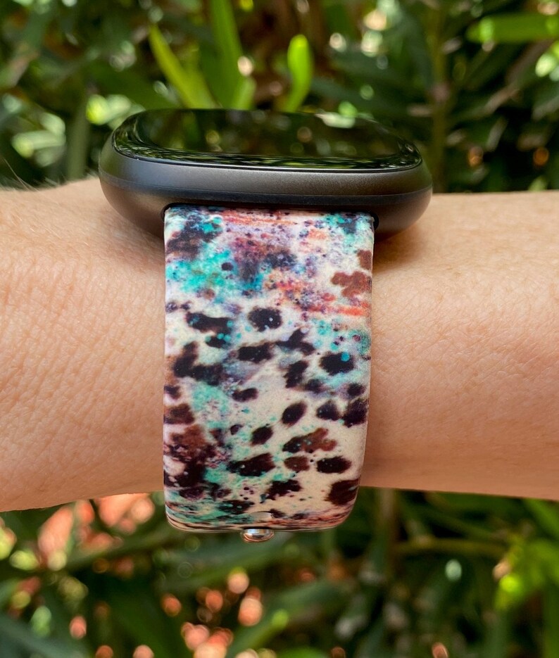 Może przedstawiać: Czarny smartwatch z kolorowym paskiem o abstrakcyjnym wzorze. Pasek ma białe tło z brązowymi, niebieskimi i turkusowymi plamami i kropkami.