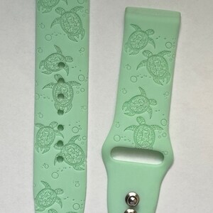 May include: A pair of light green silicone watch bands with a pattern of sea turtles and bubbles. The bands are designed for a smartwatch.