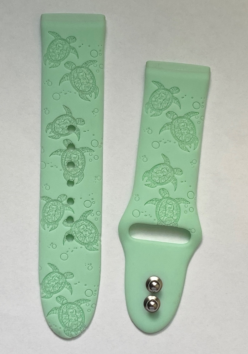 May include: A pair of light green silicone watch bands with a pattern of sea turtles and bubbles. The bands are designed for a smartwatch.