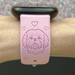 May include: A pink smartwatch band featuring a Shih Tzu dog design, hearts, and paw prints. The band is attached to a black smartwatch. The band is made of a soft, flexible material.