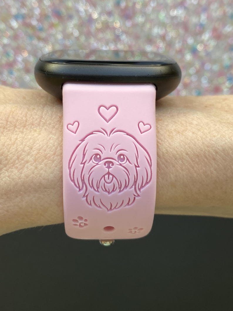 May include: A pink smartwatch band featuring a Shih Tzu dog design, hearts, and paw prints. The band is attached to a black smartwatch. The band is made of a soft, flexible material.