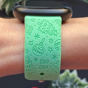 K&ouml;nnte beinhalten: Ein mintgr&uuml;nes Uhrenarmband mit festlichem Weihnachtsdesign. Das Armband zeigt gepr&auml;gte Weihnachtsb&auml;ume, Ornamente, Schneeflocken und die Worte "HO HO". Das Zifferblatt der Uhr ist schwarz.