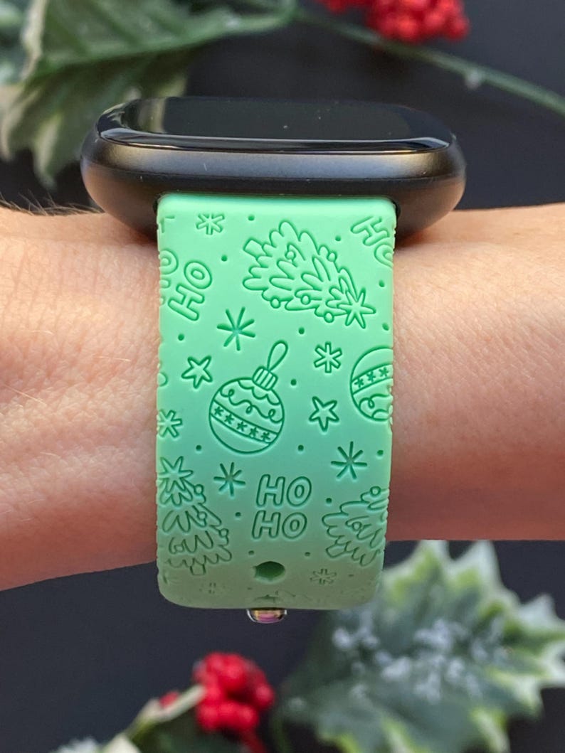 K&ouml;nnte beinhalten: Ein mintgr&uuml;nes Uhrenarmband mit festlichem Weihnachtsdesign. Das Armband zeigt gepr&auml;gte Weihnachtsb&auml;ume, Ornamente, Schneeflocken und die Worte "HO HO". Das Zifferblatt der Uhr ist schwarz.