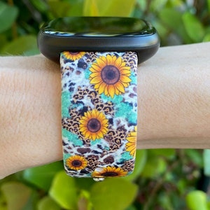 May include: A black smartwatch with a patterned band featuring yellow sunflowers, brown leopard spots, and turquoise accents. The band is attached to the watch face, showcasing a vibrant floral and animal print design.