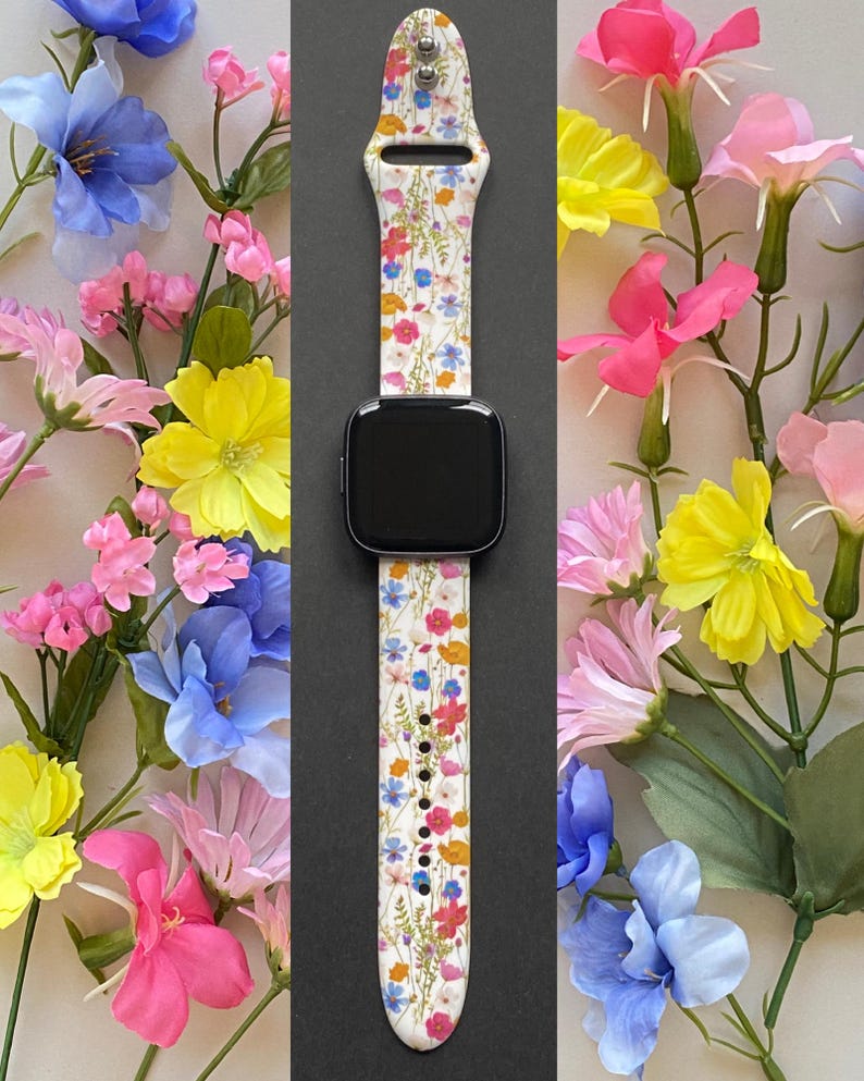 May include: A smartwatch with a white floral patterned band featuring red, blue, yellow, and orange flowers. The watch is surrounded by colorful flowers, including pink, yellow, and blue blooms, creating a vibrant display.