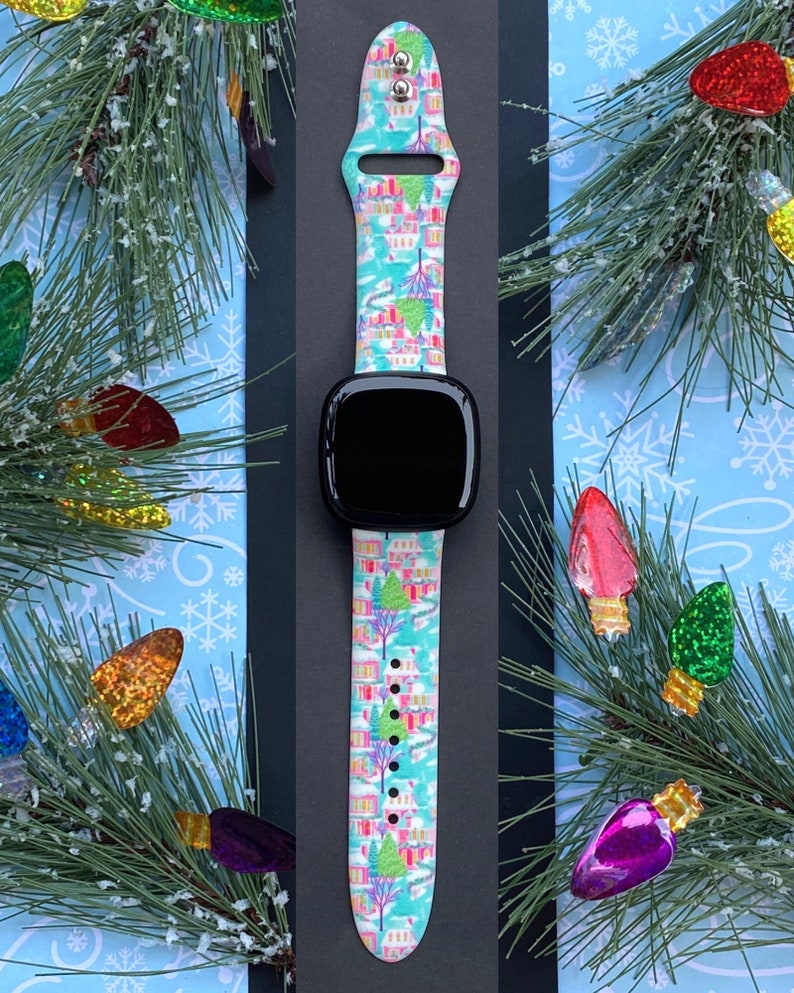 K&ouml;nnte beinhalten: Ein t&uuml;rkisfarbenes Smartwatch-Armband mit einem farbenfrohen Winterdorfmuster. Das Armband zeigt H&auml;user, B&auml;ume und Schnee mit einem schwarzen Zifferblatt. Das Armband ist mit einem Weihnachtsthema dekoriert.