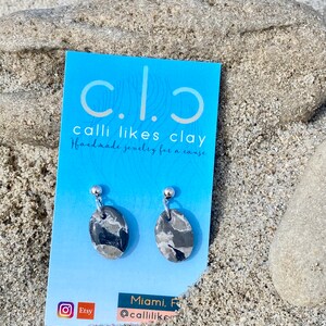 May include: A pair of gray and black oval-shaped earrings with silver posts. The earrings are on a blue card with the text "calli likes clay Handmade jewelry for a cause" and the social media handle "@callilikesclay".