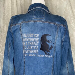 May include: A blue denim jacket with a black and white embroidered quote by Dr. Martin Luther King Jr. that reads "Injustice anywhere is a threat to justice everywhere."
