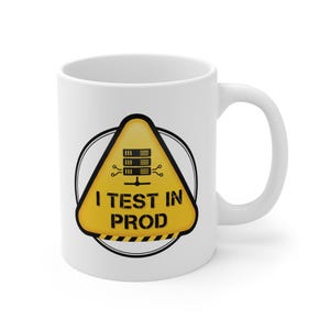 May include: White ceramic mug with a black handle. The mug features a yellow warning sign with the text "I TEST IN PROD" and a server graphic. The sign is inside a black circle.