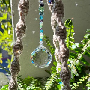 May include: A crystal suncatcher with a clear crystal prism hanging from a macrame string with blue and green beads. The suncatcher is hanging in front of a green fern plant.