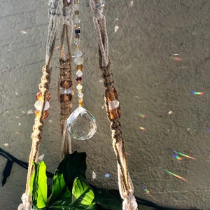 May include: A macrame plant hanger with brown and white rope, featuring a crystal prism and brown and clear beads.
