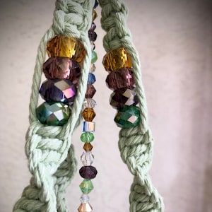 May include: A close-up of a macrame plant hanger with a beaded strand of colorful crystals. The beads are in shades of green, purple, gold, and clear.