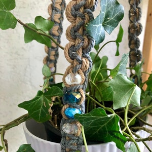 May include: A macrame plant hanger with a white pot filled with green ivy. The hanger is made of gray and tan rope, accented with clear and blue beads. The plant hanger is designed to suspend a potted plant.