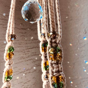 May include: A macrame wall hanging with a crystal prism and green and gold beads. The macrame is made with natural colored cord.