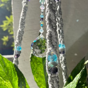 May include: A gray macrame plant hanger with crystal beads and a clear crystal prism hanging from it. The hanger is hanging from a plant.