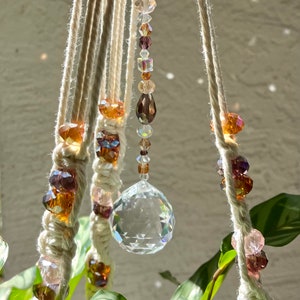 May include: A macrame plant hanger with a crystal prism and brown, clear, and pink beads. The hanger is made of natural cotton rope and is hanging from a ceiling.