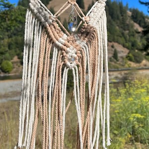 May include: A macrame wall hanging with a crystal teardrop pendant. The hanging is made with natural beige and white cotton cord and is attached to a wooden dowel.