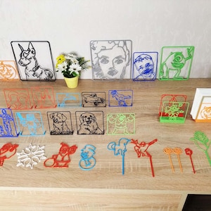 May include: A collection of 3D printed wireframe art pieces, including a dog, a monster, a pumpkin, a snowflake, a snowman, a car, a dinosaur, and a person. The pieces are all different colors and are arranged on a wooden table.