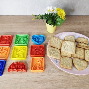 Op de afbeelding: Een set van 10 kleurrijke plastic koekjesvormen met verschillende cartoonfiguren, waaronder een hart, een hond, een vleermuis en een sneeuwpop. De vormen zijn gerangschikt op een houten oppervlak naast een bord met koekjes met bijpassende ontwerpen.