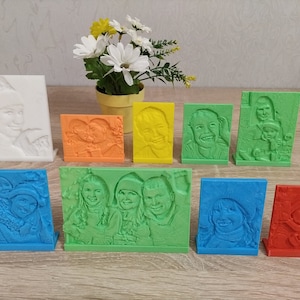 Puede incluir: Una colección de 10 pequeños marcos de fotos impresos en 3D, coloridos, con diferentes imágenes de personas. Los marcos están dispuestos en dos filas sobre una superficie de madera.