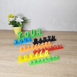 Puede incluir: Un conjunto de siete placas de nombres de colores, cada una con un nombre diferente escrito en letras 3D. Los nombres son "YOUR", "YOUR", "DARTH", "SKYWALKER", "DONALD", "JIM" y "SHERLOCK".