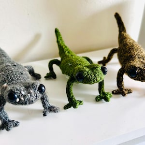 May include: Three handmade crochet geckos in gray, green, and brown. Each gecko has large black eyes and is posed with its legs extended. The geckos are displayed on a white surface, showcasing their detailed craftsmanship.