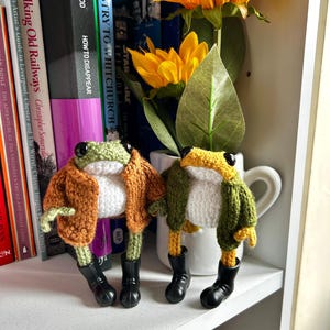 May include: Two handmade crochet frog figurines. One frog wears an orange jacket, the other a green jacket. Both have white bellies, green heads, and black boots. They stand on a white shelf next to a vase of yellow flowers and books.