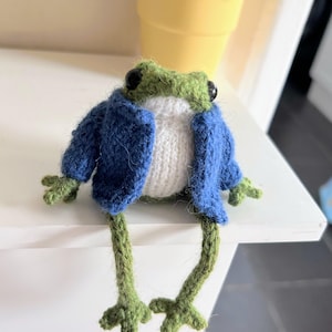May include: A knitted frog toy, featuring a green head and legs, a white body, and a blue jacket. The frog has black button eyes. It is sitting on a white surface. A yellow plant pot is visible in the background.