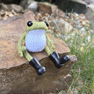 May include: A small, handmade crochet frog figurine featuring a light green head and limbs with a white belly. The frog has large, round, black plastic eyes and is wearing a pair of miniature, solid black rubber rain boots on its feet.
