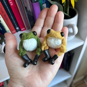 May include: Two small needle-felted frogs, one green and one yellow, each featuring a white belly, large black plastic eyes, and miniature black rubber boots. The frogs are crafted from textured wool fibers and are positioned side by side.