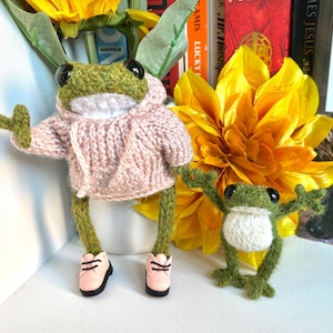 May include: Two handmade crocheted green frogs with black eyes and white bellies. The larger frog wears a light pink knitted hoodie and matching pink shoes. The smaller frog stands with arms raised. Both have long, thin limbs and textured green yarn bodies.