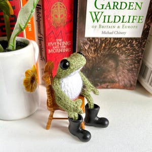May include: A small, crocheted green frog with a white belly and large black eyes, wearing black plastic boots. The frog is seated on a miniature wooden chair. The frog has textured green yarn limbs and a rounded head, positioned upright on the chair.