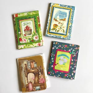 May include: Three rectangular notebooks with decorative covers. One features a green floral pattern with a basket of strawberries and the text "Life is Sweet". Another has a blue floral pattern with a landscape and "Map Your Memories". The third is brown with a vintage sewing machine illustration.